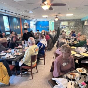 A large group of people sit at round tables in Café Lee, eating, talking, and socializing. The lively atmosphere features bright lighting, modern decor with blue and brick walls, ceiling fans, and a buzz of student leadership energy.
