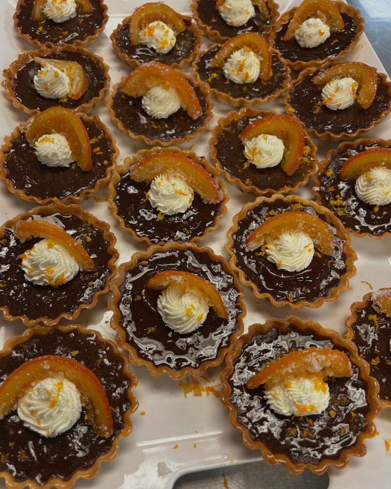 Mini chocolate tarts from Café Lee are topped with a swirl of whipped cream and a slice of candied orange, arranged closely together on a white tray—perfect for student leadership events or casual gatherings alike.