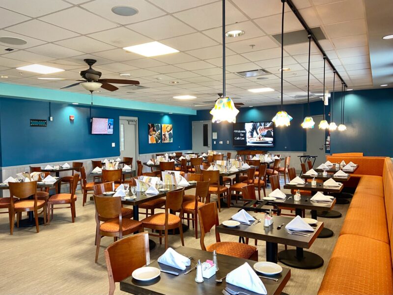 A modern restaurant dining area with neatly set tables, orange cushioned chairs, blue walls, and hanging pendant lights offers a bright, inviting space. Café Lee is perfect for gatherings or quiet moments—returns are always met with warmth.