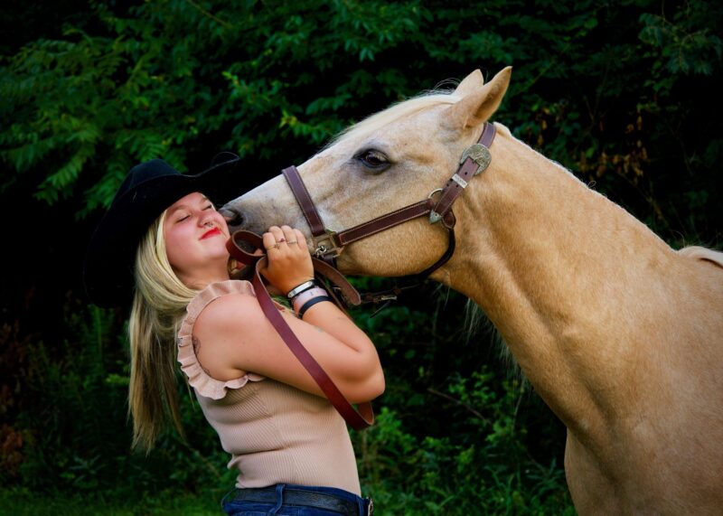 A young woman wearing a black cowboy hat smiles and scrunches her face as Kacee the Wonder Pony nuzzles her cheek in a green outdoor setting.