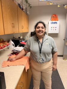 A woman wearing a gray polo and khaki pants stands smiling in a break room with wooden cabinets, showing empathy as she rests her hand on folded clothes for the Care Closet. Snacks and cleaning supplies are visible nearby.