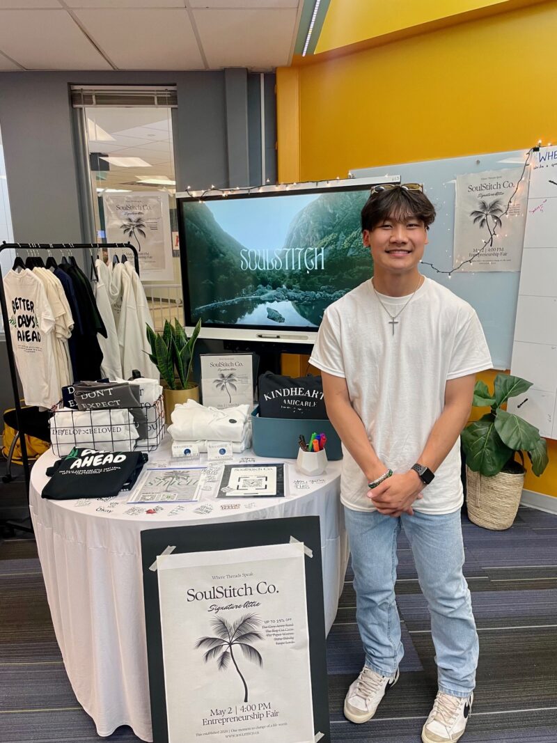 A person stands smiling in front of a SoulStitch Co. booth displaying apparel, prints, and a screen with the brand’s logo at an indoor event—an inspiring scene of entrepreneurship and creativity. Posters and T-shirts are arranged on tables and racks nearby.