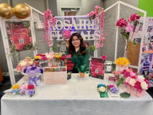 A woman with long dark hair and glasses smiles behind a table displaying colorful artificial flowers, showcasing her entrepreneurship at "Rosaire Florals." The booth features innovative floral arrangements with pink and gold accents.