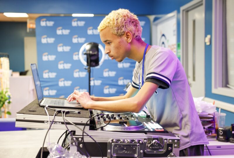 A person with short blond hair operates DJ equipment and a laptop in a brightly lit room with blue walls and a step-and-repeat banner, capturing the energy of entrepreneurship and building the future.