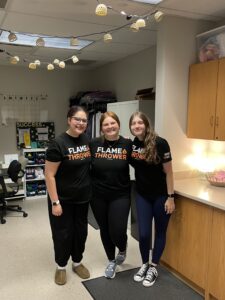 Three women stand together smiling in a classroom, all wearing matching black “Flame Thrower” T-shirts. Cabinets, desks, and string lights decorate the room around them, reflecting their shared impact through initiatives like the Care Closet.