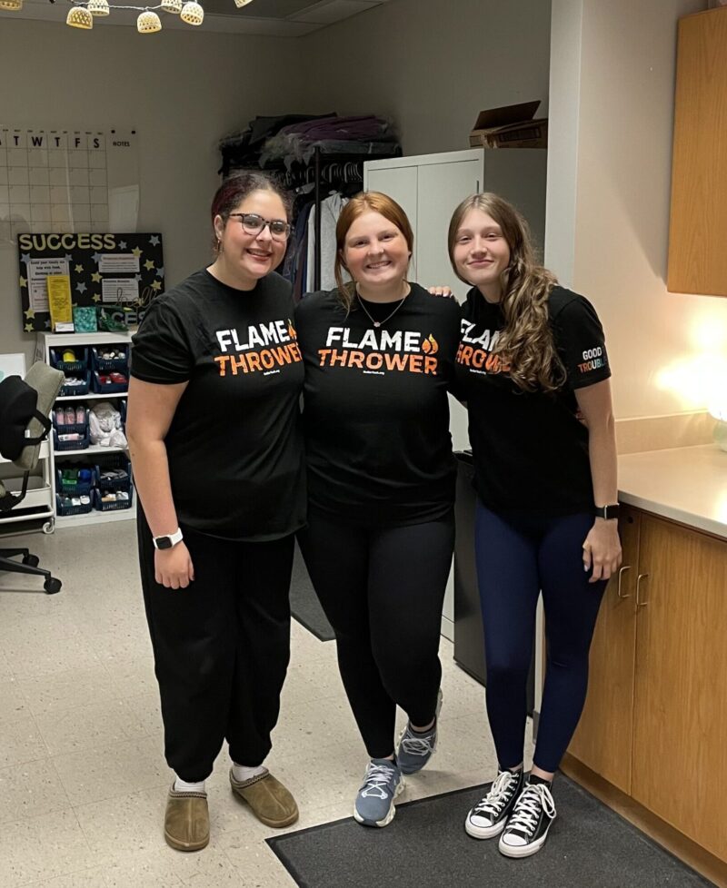 Three women stand side by side in a classroom, smiling at the camera and wearing matching black 