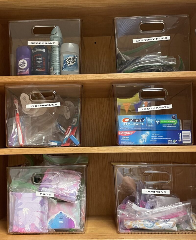 Clear plastic bins on wooden shelves, each labeled and containing hygiene items like deodorant, laundry pods, toothbrushes, and more—all neatly organized in the Care Closet to support the community with empathy and impact.