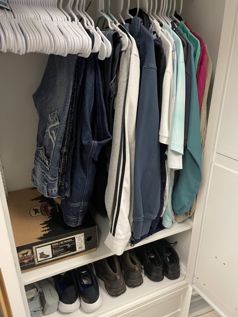 A Care Closet with shirts, jackets, and denim vests on white hangers, plus shoes and a shoebox neatly on the lower shelf. This organized, white wardrobe reflects empathy through its thoughtful arrangement.