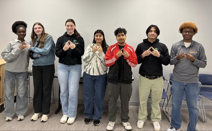 Seven business students stand side by side indoors, facing the camera and making triangular hand signs. They wear casual clothing and smile in a room with neutral walls and chairs, representing their DECA chapter.