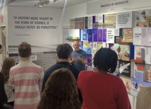A group of people listen to a man speaking in front of a display wall about innovation and entrepreneurship. A Kipling quote about history and stories is visible on the wall, with various posters and images highlighting the Year in Review.