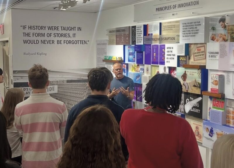A group of people listen to a man speaking in front of a display wall about innovation and entrepreneurship. A Kipling quote about history and stories is visible on the wall, with various posters and images highlighting the Year in Review.