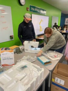 Two people work together at a table with a sewing machine and various materials, showcasing hands-on entrepreneurship. One operates the machine while the other observes. The room features green walls, a "Small Business" sign, and boxes of tools on the table.