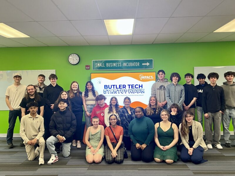 A group of young adults pose and smile in front of a screen that reads “Butler Tech Startup Week” in a bright room with green walls, celebrating entrepreneurship. A sign above points to a “Small Business Incubator.”.