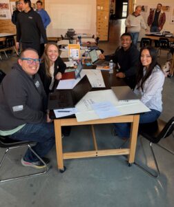 Four people sit around a table with laptops and papers, smiling at the camera in a classroom or workshop focused on entrepreneurship. Other groups work in the background, and the wood-paneled room suggests an inspiring education setting.