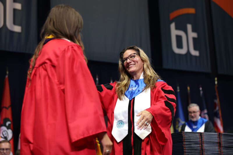 A woman in academic regalia, representing Human Resources, smiles and congratulates a graduate in a red gown on stage at a graduation ceremony, with banners and flags visible in the background.