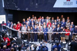 A large group of people, dressed mostly in business attire, stand on stage holding a ribbon at a Miami University ribbon-cutting ceremony, as photographers capture the event—signaling that the work continues beyond this milestone.