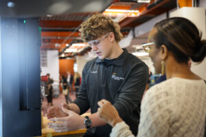 A young man wearing safety goggles and a dark shirt gestures with his hands while talking to a woman with glasses and a patterned sweater beneath bright lights, as work continues in the modern workspace.
