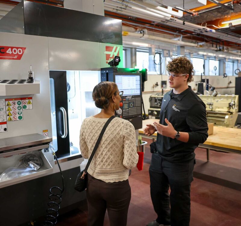 A man explains how to operate a UMC 400 CNC machine to a woman in a busy workshop, where work continues and other equipment is visible in the background.