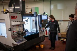 A person demonstrates how to operate a CNC machine labeled VF2 in a workshop, with several people observing as work continues. The machine features a control panel, various safety labels, and a prominent ribbon marking its recent installation.