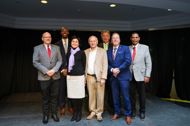 Seven people stand on a stage in formal attire, posing for a group photo in front of black curtains. A ribbon is visible nearby, hinting at a recent ceremony, but the work continues as they celebrate this achievement together.