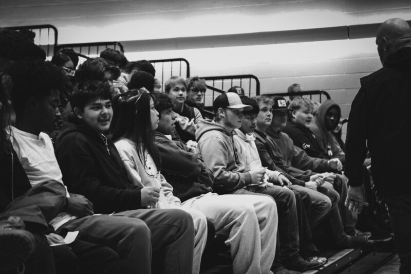 A group of teenagers sit closely together on bleachers in a gymnasium during 2026 Apprenticeship Day, wearing casual clothes and hoodies. An adult stands in front of them, and the mood appears casual and attentive. The image is in black and white.