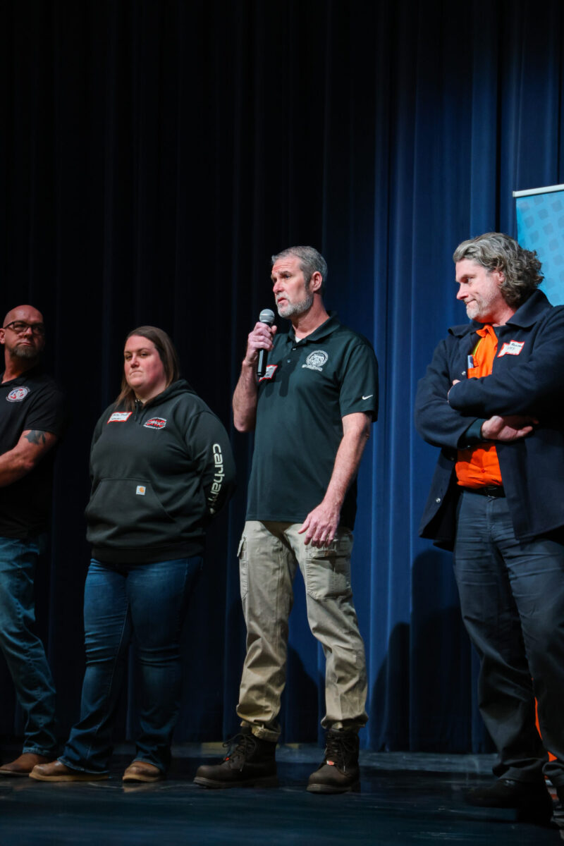 Four people stand on stage in front of blue curtains. The man in the center speaks into a microphone. Each person wears casual or work clothing and looks attentive at the 2026 Apprenticeship Day event.