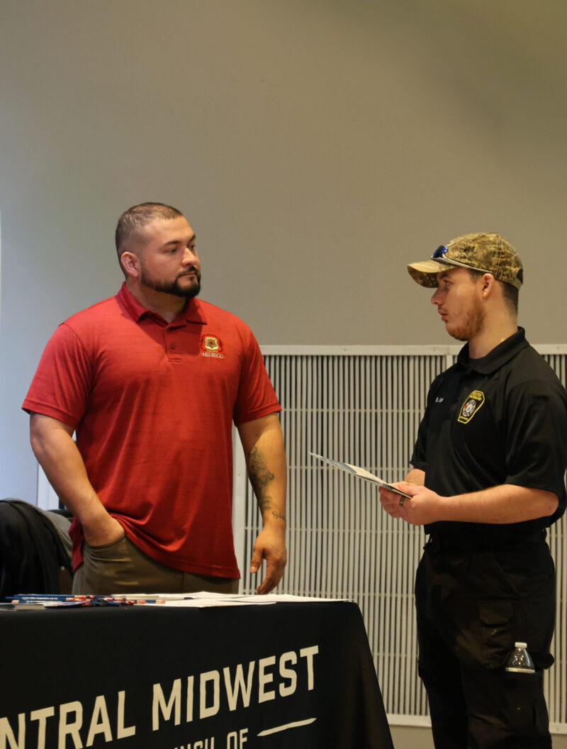 Two men talk at a table, one in a red polo and one in a black security uniform with a camo hat, holding papers. The table displays 