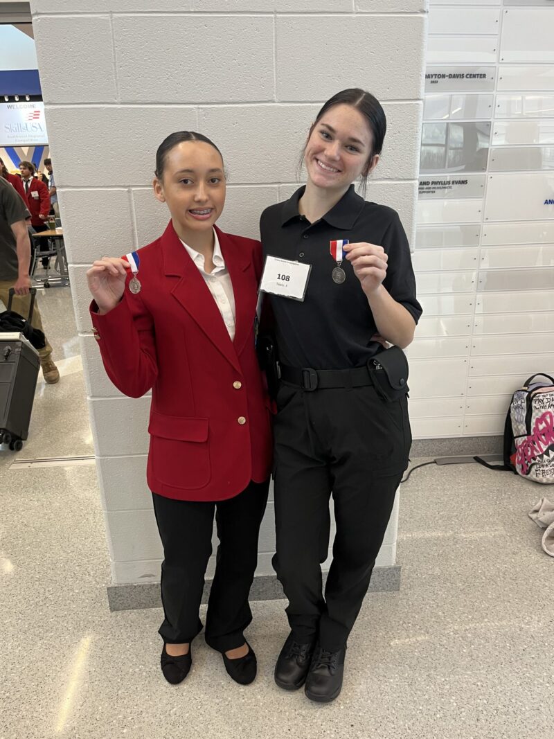 Two young women stand indoors, smiling and holding medals. One wears a red blazer while the other, a BT student in a black uniform with a contest number tag, celebrates their success at a SkillsUSA Regional Competition event.