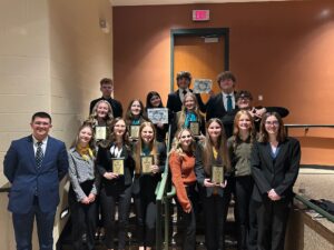 A group of students in business attire pose indoors on stairs and a landing, smiling and holding awards. Two adults stand at either end of the group. The background features a tan wall and a closed door.