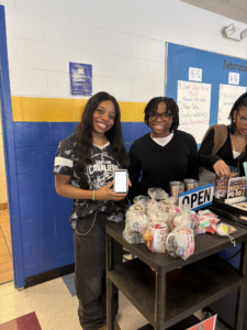 Two smiling students stand behind a DECA School-Based Enterprise snack cart filled with treats. One holds up a smartphone displaying a screen. The cart has an "OPEN" sign, mugs, and donation box, with posters on the blue and yellow wall behind them.