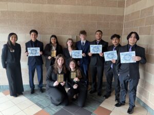 A group of nine students in business attire pose indoors with certificates and plaques, smiling. They stand in front of a tiled wall, celebrating their achievements, with one adult standing on the left.