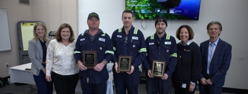 Eight people pose indoors; three apprentices in work uniforms hold plaques, while five in business attire stand on either side. A large monitor and papers are visible, suggesting an award or recognition event celebrating careers.