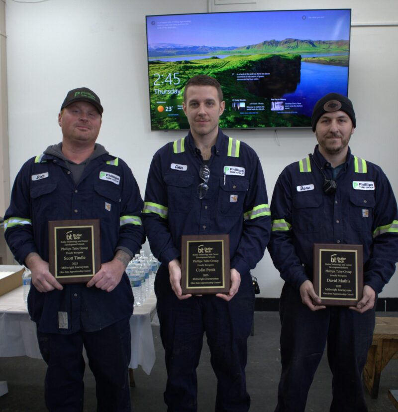 Three men in blue work uniforms stand side by side indoors, each holding a wooden plaque. Behind them, a TV displays a landscape and current time. A table with water bottles is visible to the left.