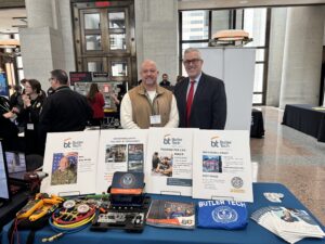 Two men stand behind a Butler Tech informational booth featuring CTE program displays, photos, pamphlets, and colorful tools. The indoor event at the Ohio Statehouse includes other attendees in the background.