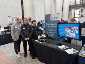 Four BT students stand next to a table showcasing a robotics project at the Butler Tech engineering booth inside the Ohio Statehouse. The display features technology-related signs, brochures, a monitor, and a wheeled robot model.