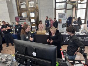 A group of people stand around a booth at a technology expo in a large hall, with informational displays, computers, and robotics equipment visible. Two presenters speak with an attendee near the center, highlighting opportunities for BT Students in CTE programs.
