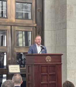 A man stands at a podium giving a speech inside the Ohio Statehouse, as several people, including BT students, sit and listen. An American and state flag are visible to the right of the podium.