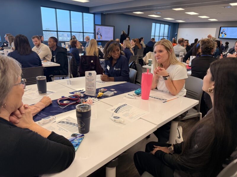 A group of people sit around tables in a bright, modern conference room, engaged in lively discussion about future opportunities. Papers, pens, and drinks are on the tables as others network and form partnerships in the background.