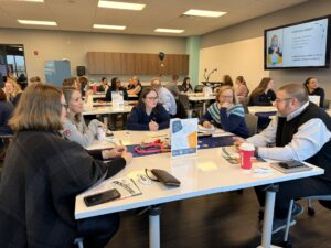 A group of people sit at tables in a brightly lit meeting room, engaged in discussion. Papers, drinks, and materials are on the tables as the Business Advisory Council explores partnerships and future opportunities, with a presentation displayed on a screen.