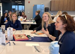 Four women sit around a table in a bright, modern meeting room, smiling and talking about future opportunities. Papers, flyers, and a blue Butler Tech water bottle are on the table, with more people networking in the background.