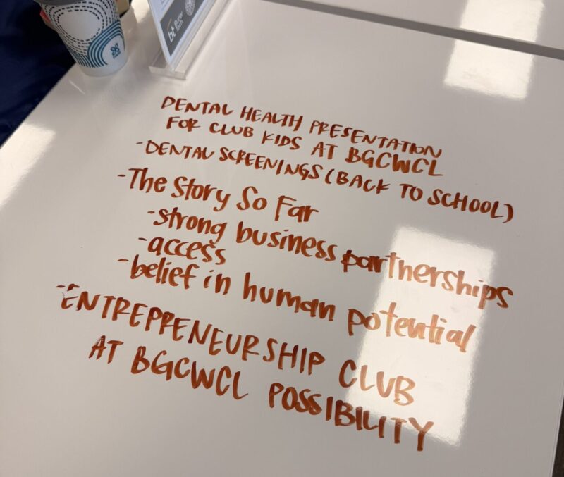 A whiteboard with handwritten notes in orange marker about a dental health presentation and entrepreneurship club for BGCGWCL kids, mentioning dental screenings, partnerships, access, future opportunities, and belief in human potential. A coffee cup is nearby.