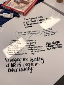 A tabletop with handwritten notes about healthcare, a quote on quality of life in Butler County, a philosophy book, and a magazine. The notes highlight inclusivity, education, healthcare impact, and future opportunities for partnerships.