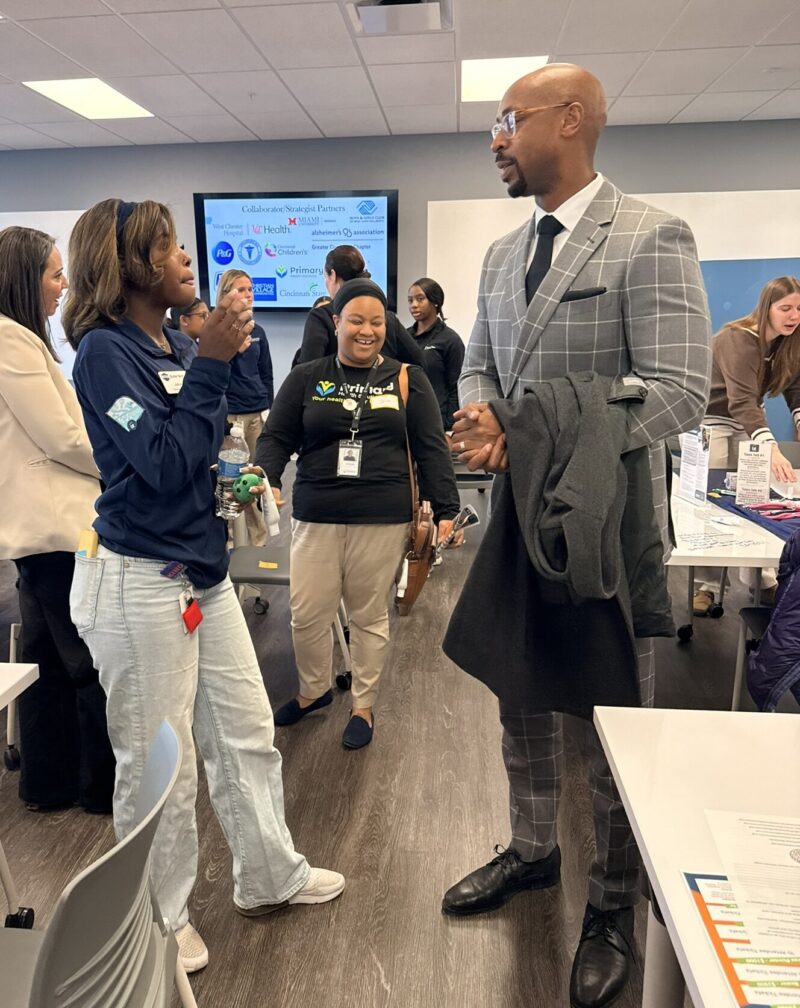 Several professionals, some in business attire and others in medical scrubs, share ideas and engage in a bright, modern conference room with tables, informational materials, and a presentation screen highlighting key partnerships.
