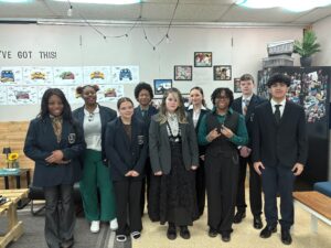 A group of nine high school students dressed in business attire stand together in a classroom, smiling at the camera. The classroom walls have posters and photos, and there’s a cozy seating area to the side.