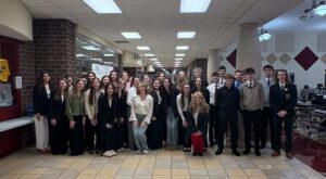 A group of young adults dressed in formal attire stand closely together in a school hallway with a tiled floor and fluorescent lighting, smiling at the camera.