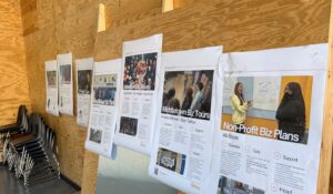 Several large printed posters with business-related and entrepreneurship content are displayed on a wooden board in a modern room. Stacks of black chairs are visible to the side, suggesting an office or education-focused workshop space.
