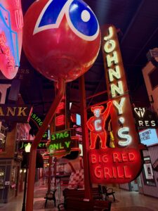A vibrant indoor display at the American Sign Museum showcases vintage neon signs, including a large red sphere and "Johnny’s Big Red Grill," illuminating a retro-themed walkway with benches and sparking inspiration in entrepreneurship.