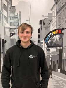 A young person with light brown hair, wearing a black "Automotive Technology" hoodie, stands on campus in front of a mural of a city street with a colorful "EMPOWER" sign overhead, ready to gain valuable automotive experience.