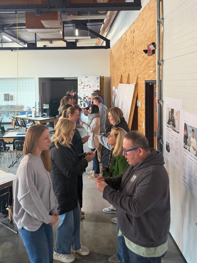 A group of people stand in two lines facing each other inside a modern workspace, engaging in conversation about entrepreneurship. Some hold papers or phones. The room features wooden and white walls, tables, and artwork displayed.