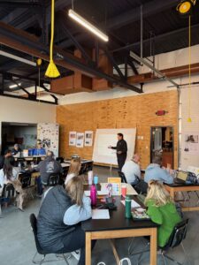 A group of adults sits at tables in a classroom, focused on a presenter by a whiteboard. The room, filled with natural light and posters on a wooden wall, fosters education and entrepreneurship in an inspiring setting.